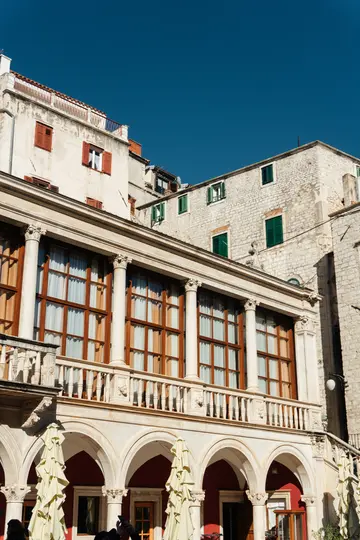 A multi-story building with a large balcony supported by columns and arches, under a clear blue sky. The building features…