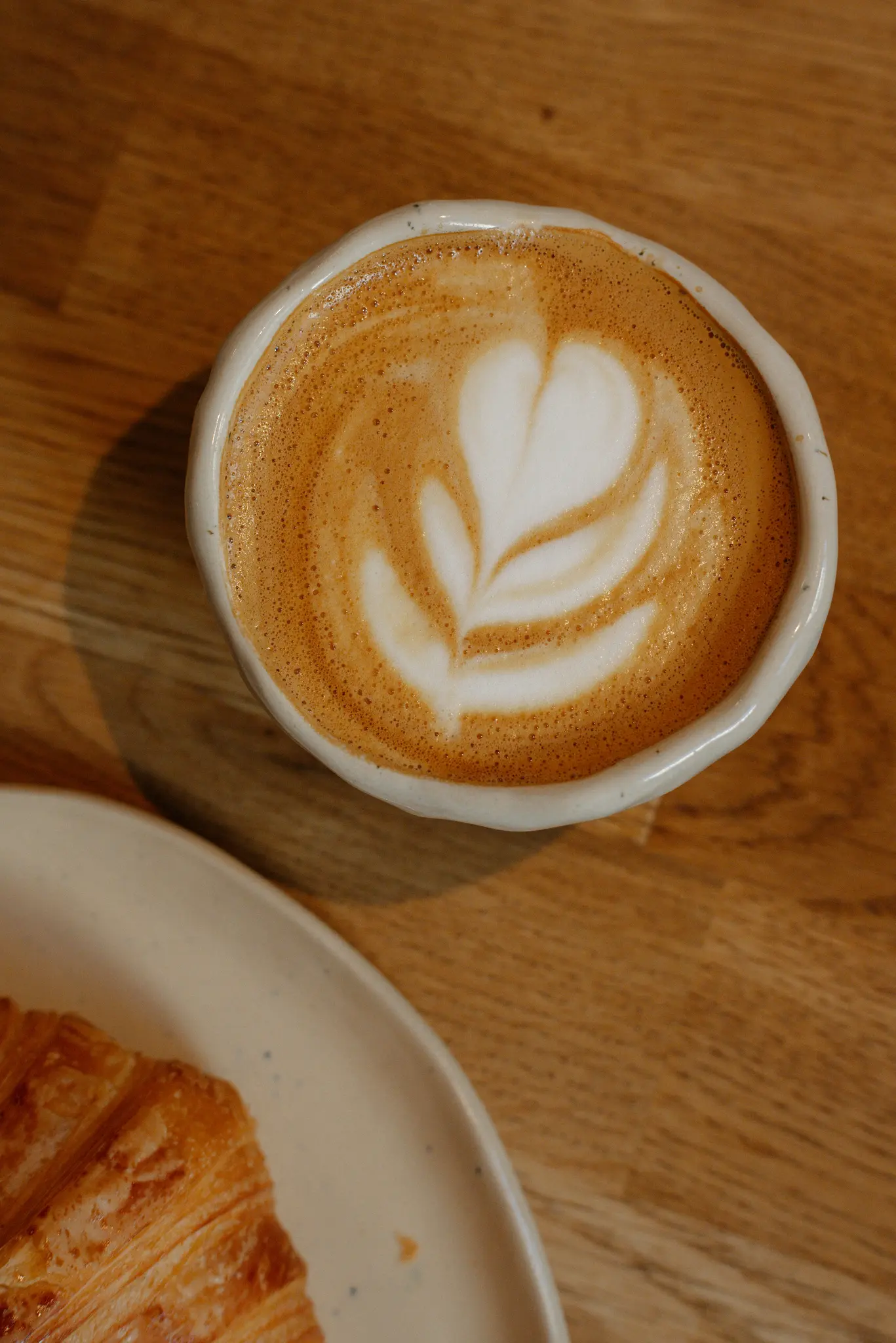 Un primo piano dall'alto di una tazza in ceramica bianca con latte art e un disegno a forma di cuore, accanto a un croissant