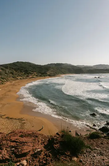 Een luchtfoto van een breed zandstrand met schuimende golven die vanaf de oceaan binnenrollen, geflankeerd door weelderige