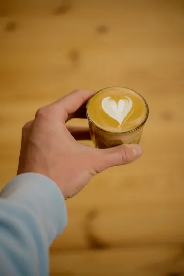 A person's hand, wearing a light blue sleeve, holds a small clear glass of latte with a white heart design in the foam.