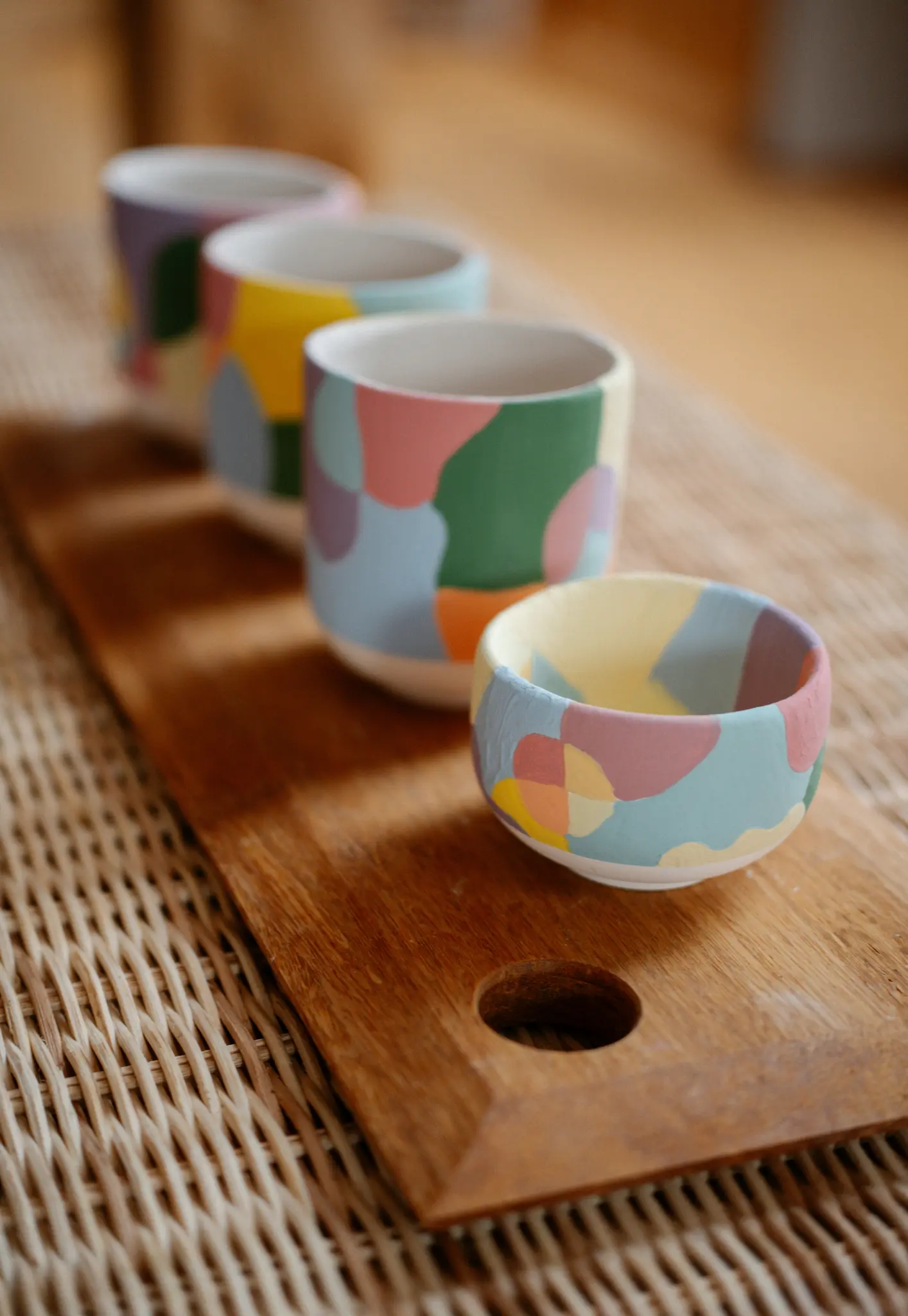 Quatre récipients en céramique, trois tasses et un petit bol, avec des motifs abstraits pastel, sont disposés sur une planche