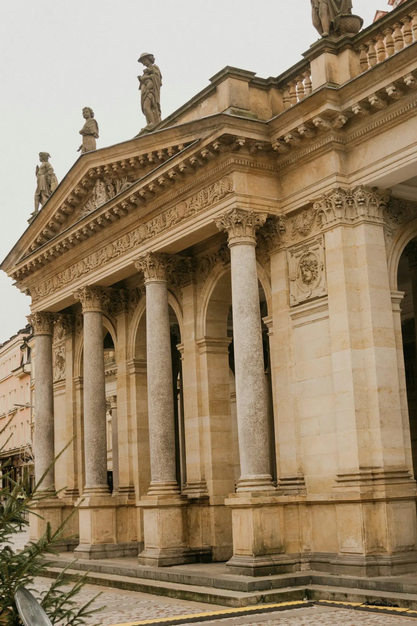 Un grande edificio in pietra chiara con più colonne alte, archi e intagli intricati. Tre statue si ergono su...