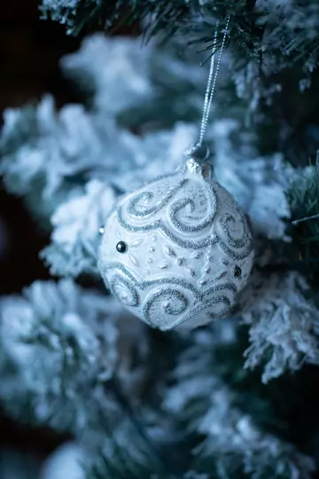 A close-up shot of a white Christmas ornament with silver glitter swirls and small black beads, hanging on a frosted…