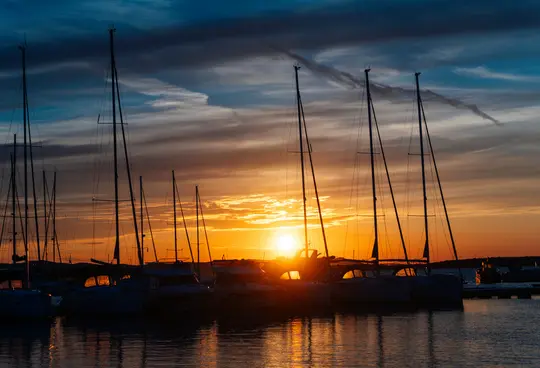 A wide landscape photo of a marina at sunset, with numerous sailboats silhouetted against a bright orange and yellow sky.