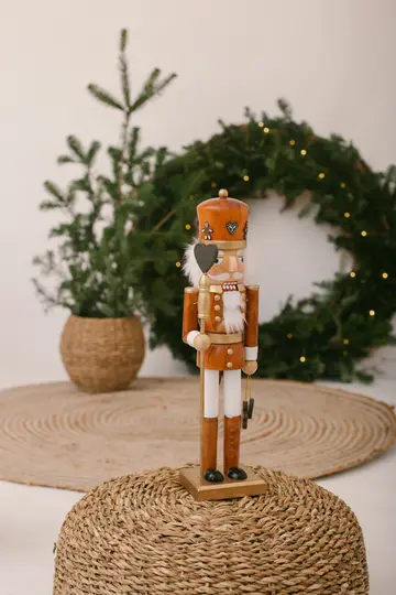 A wooden nutcracker figure in a brown and white uniform stands on a woven mat. In the background, a Christmas wreath with…