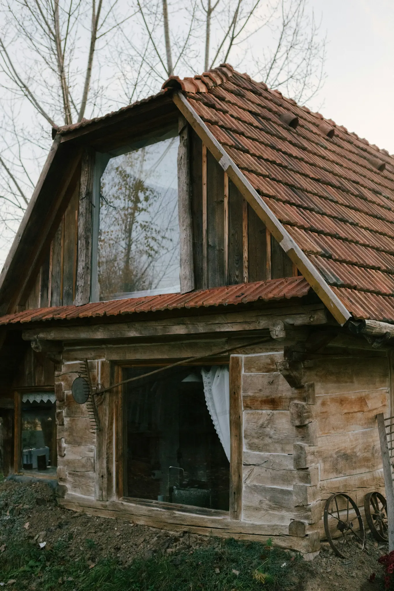 Eine alte Holzblockhütte mit rotem Ziegeldach und großen Fenstern, die kahle Bäume widerspiegeln, steht in ländlicher