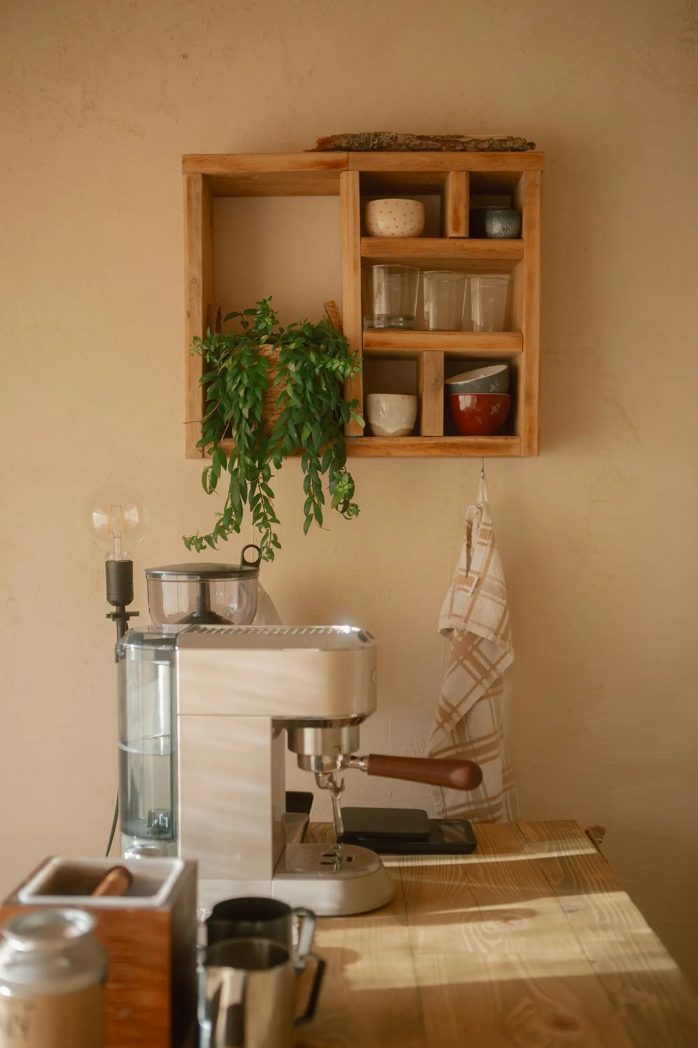 Un acogedor rincón de café con una máquina de espresso de color claro, un estante de madera con cuencos y vasos, y una planta