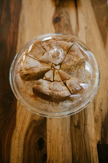 An overhead shot of a round cake, cut into multiple slices, resting on a clear glass cake stand with a dome lid. The cake…