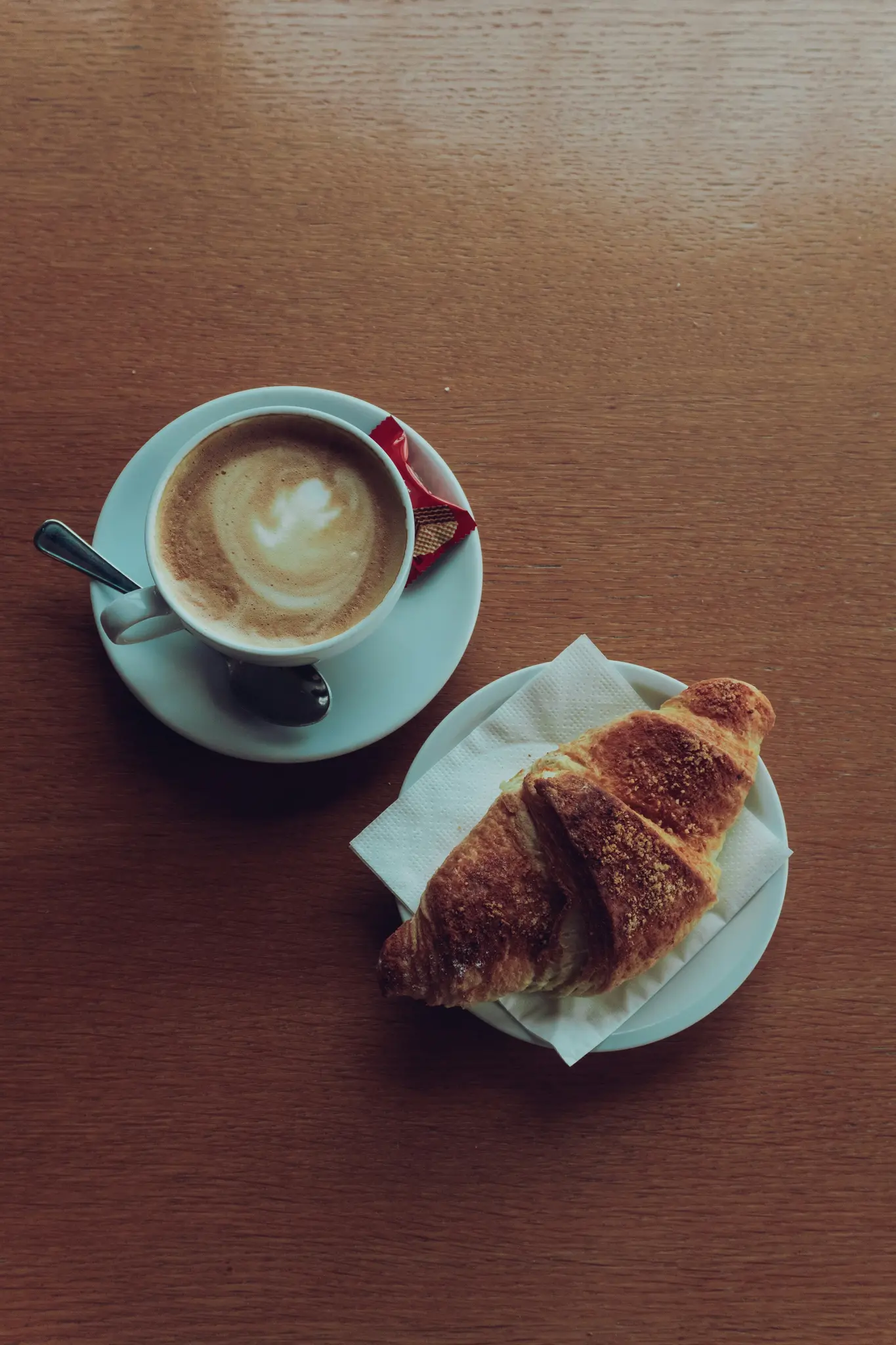 Bovenaanzicht van een witte kop koffie met latte art en een lepeltje op een schoteltje, naast een croissant op een wit bord