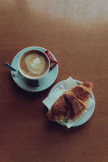 Vista dall'alto di una tazza di caffè bianca con latte art e un cucchiaino su piattino, accanto a un croissant su un piatto