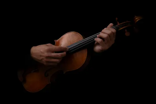 Una toma de primer plano muestra dos manos tocando un violín de madera marrón sobre un fondo completamente negro. Las manos