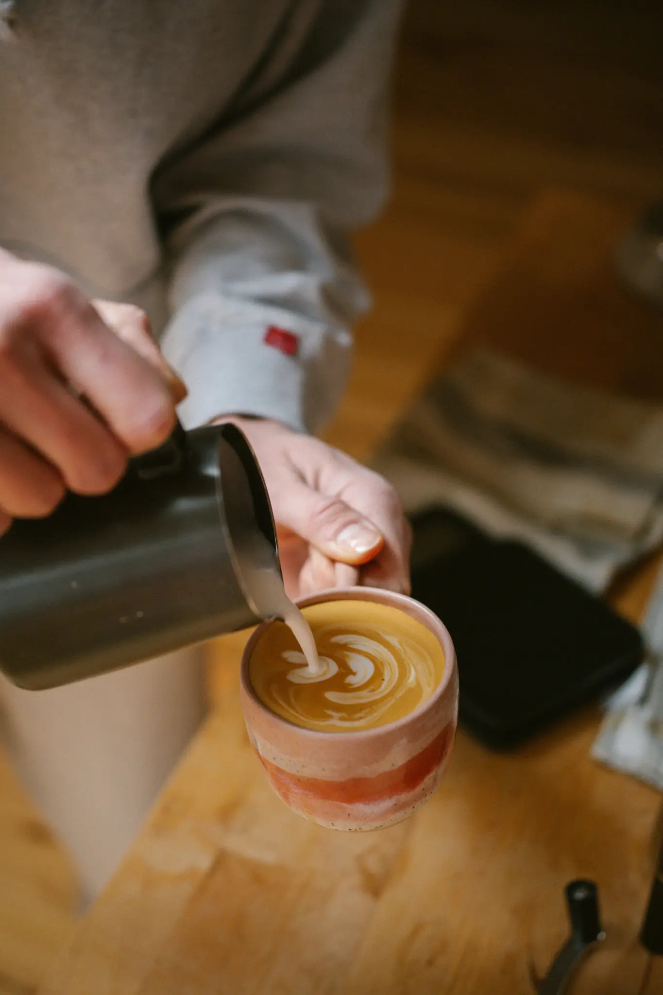 Gros plan sur des mains versant du lait mousseux blanc d'un pichet foncé dans une tasse à café en céramique, créant du latte