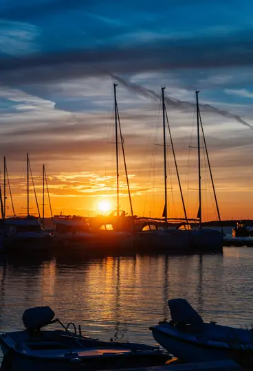 Dramatičan zalazak sunca nad marinom, sunce nisko na horizontu baca narančasti sjaj. Vide se jedrilice i motorni čamci.