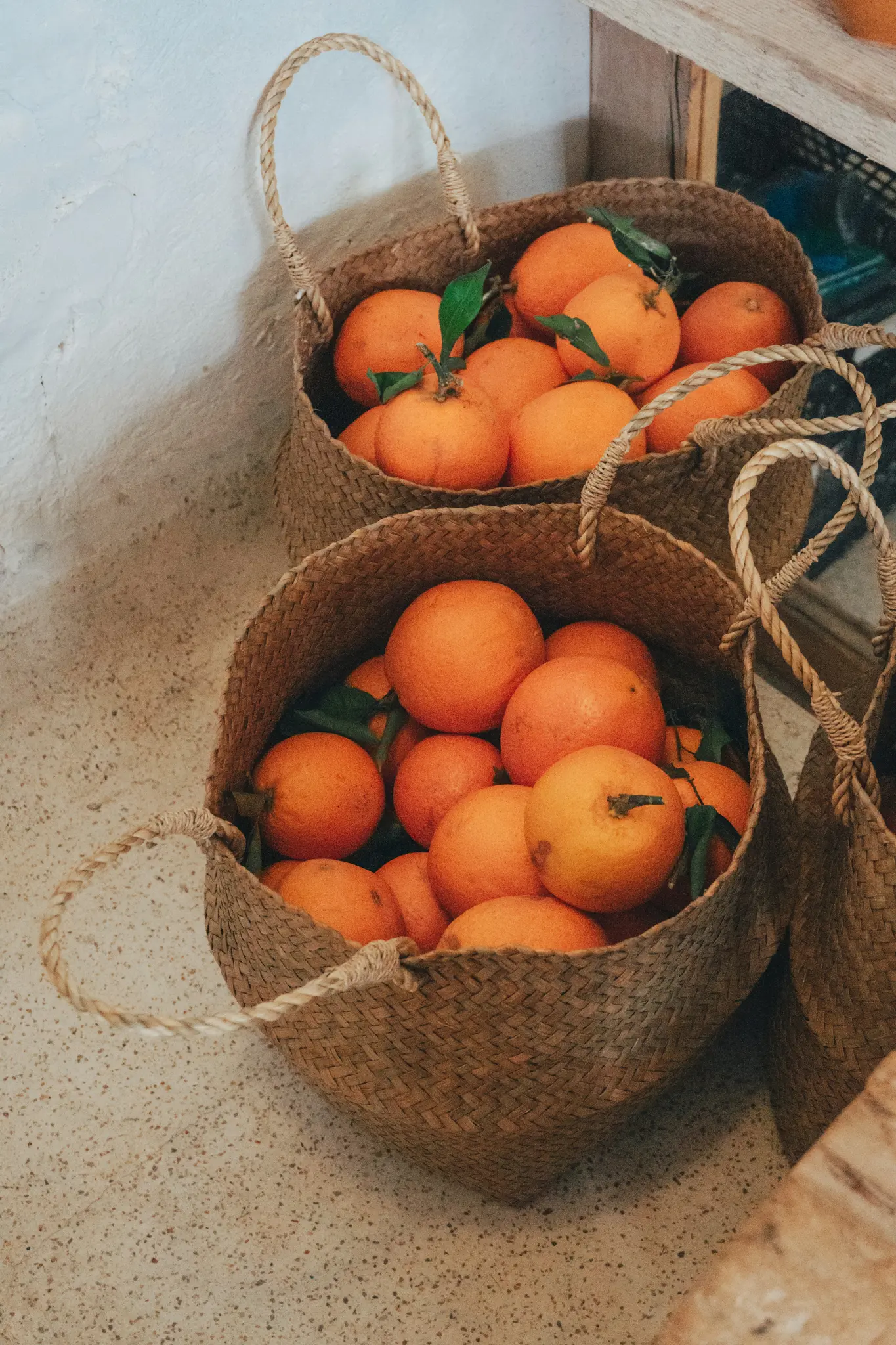 Drie geweven manden vol feloranje sinaasappels, sommige met groene bladeren, staan op een gespikkelde vloer naast een witte…