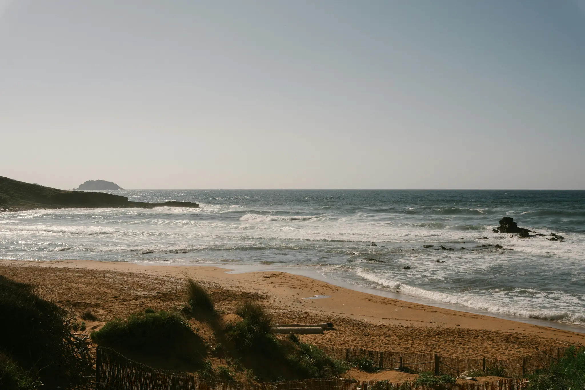 Een brede landschapsfoto van een zandstrand met zachte golven die op de kust rollen. Een klein, rotsachtig eiland is in de