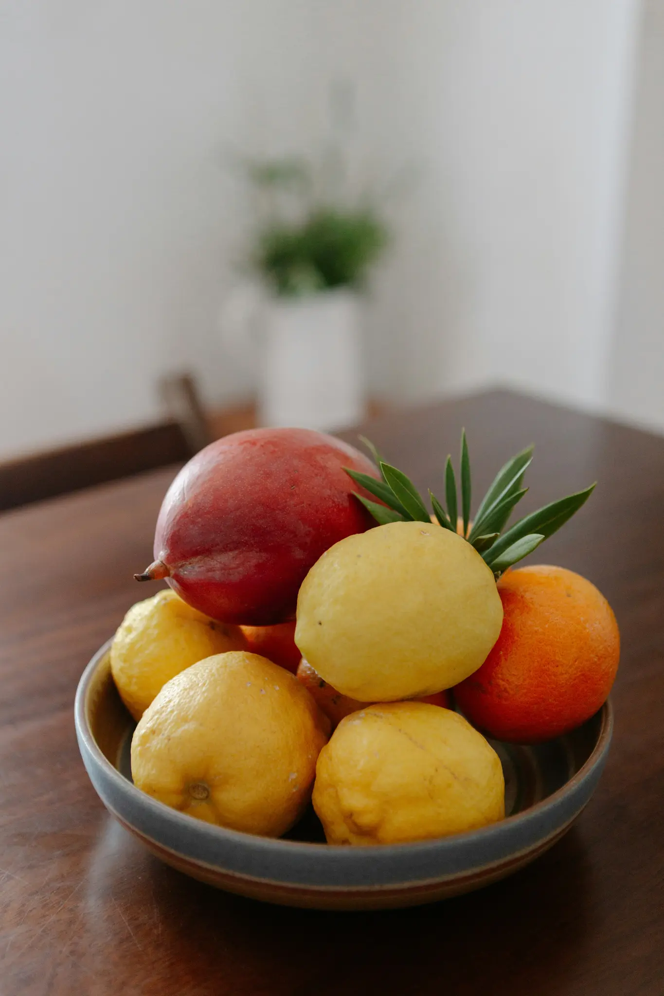 Una ciotola di ceramica piena di limoni, un'arancia e un frutto rosso, forse un mango, si trova su un tavolo di legno scuro.