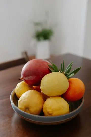 A ceramic bowl filled with lemons, an orange, and a red fruit, possibly a mango, sits on a dark wooden table. Green leaves…
