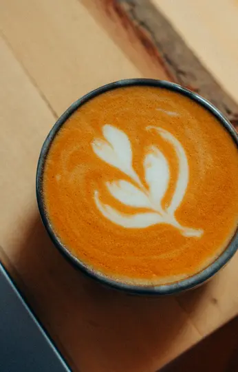 Primo piano di un latte in una tazza con bordo scuro, con arte di foglia bianca sulla schiuma su una superficie arancione.