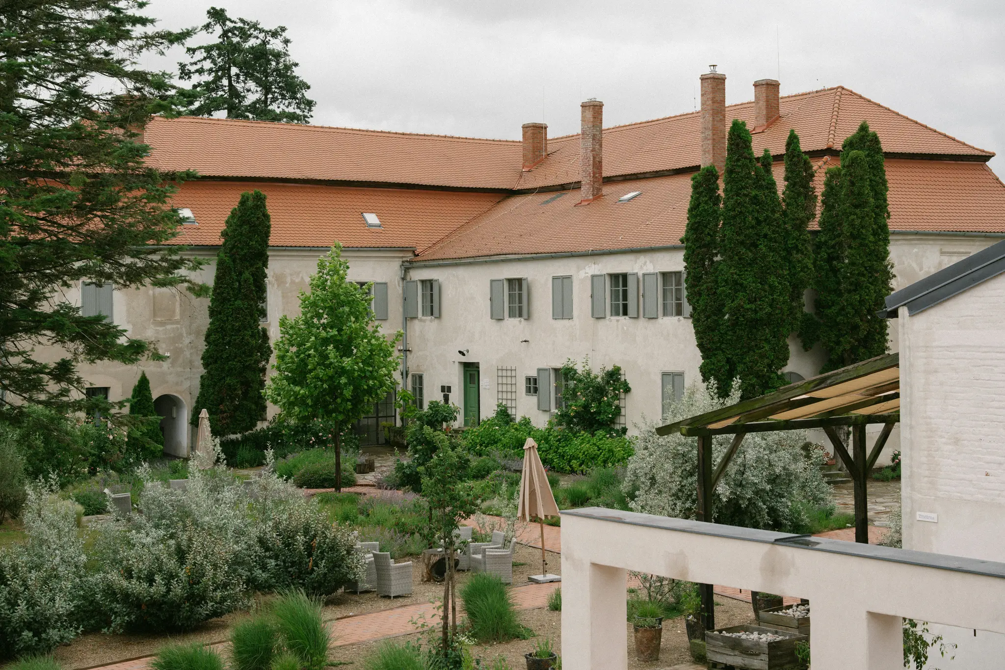 Un grande edificio di colore chiaro con tetto in tegole di terracotta e più camini si affaccia su un lussureggiante giardino