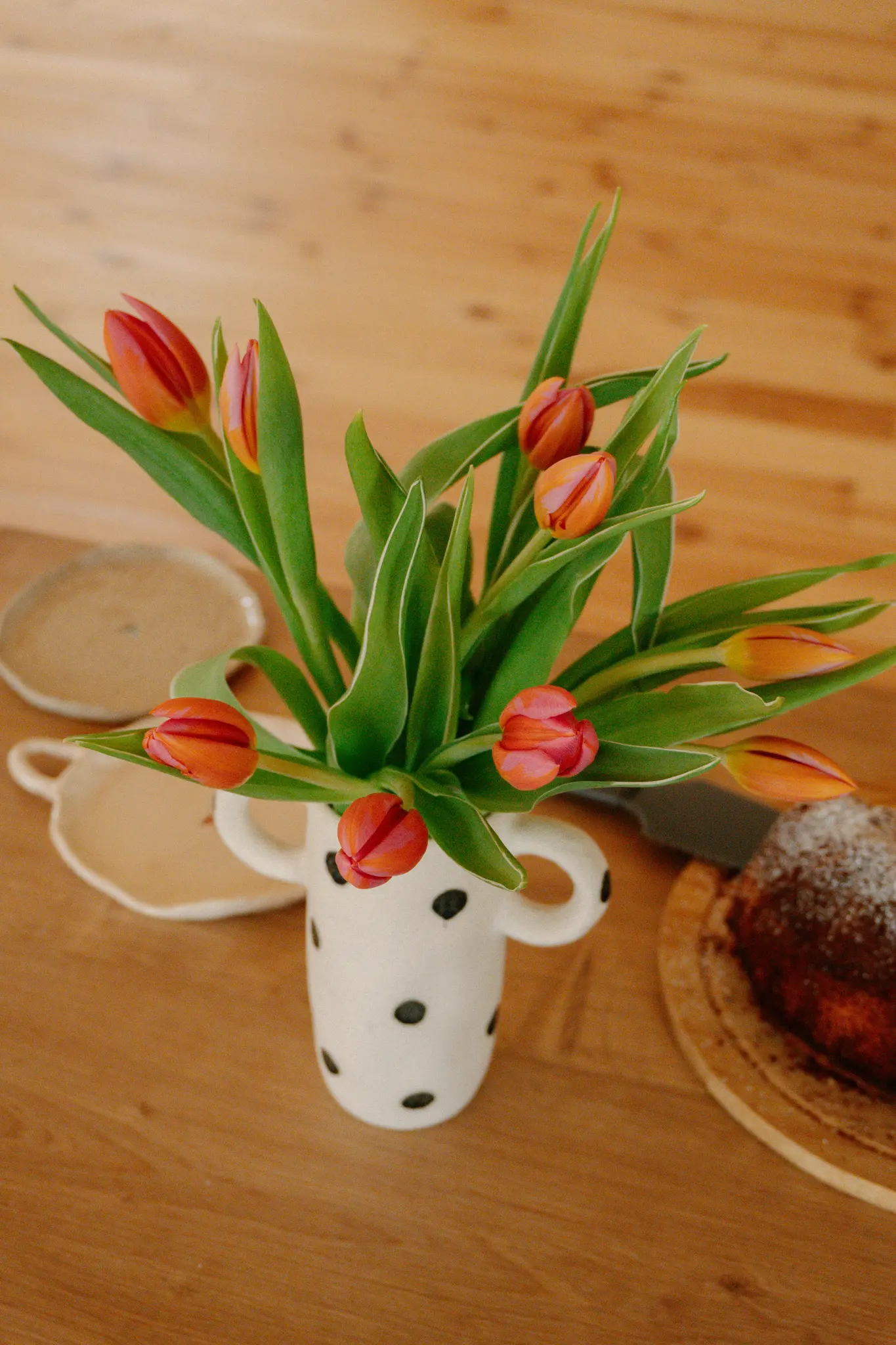 Een witte vaas met zwarte stippen bevat een boeket oranje en roze tulpen op een houten tafel. Daarnaast staan twee keramische