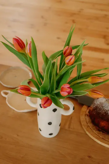 Bílá váza s černými puntíky drží kytici oranžových a růžových tulipánů na dřevěném stole. Vedle jsou dva keramické talíře a