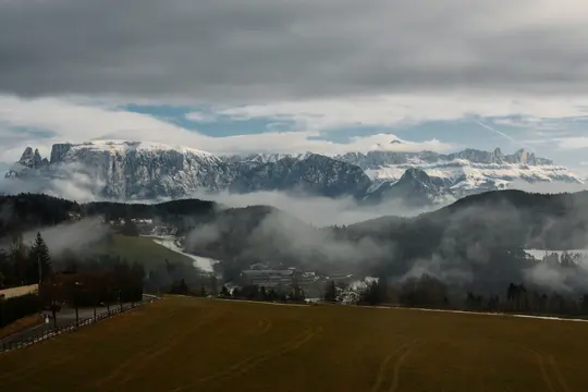 Široká krajinářská fotografie zobrazující zelené pole v popředí, údolí s mlhou a stromy a zasněžené hory.