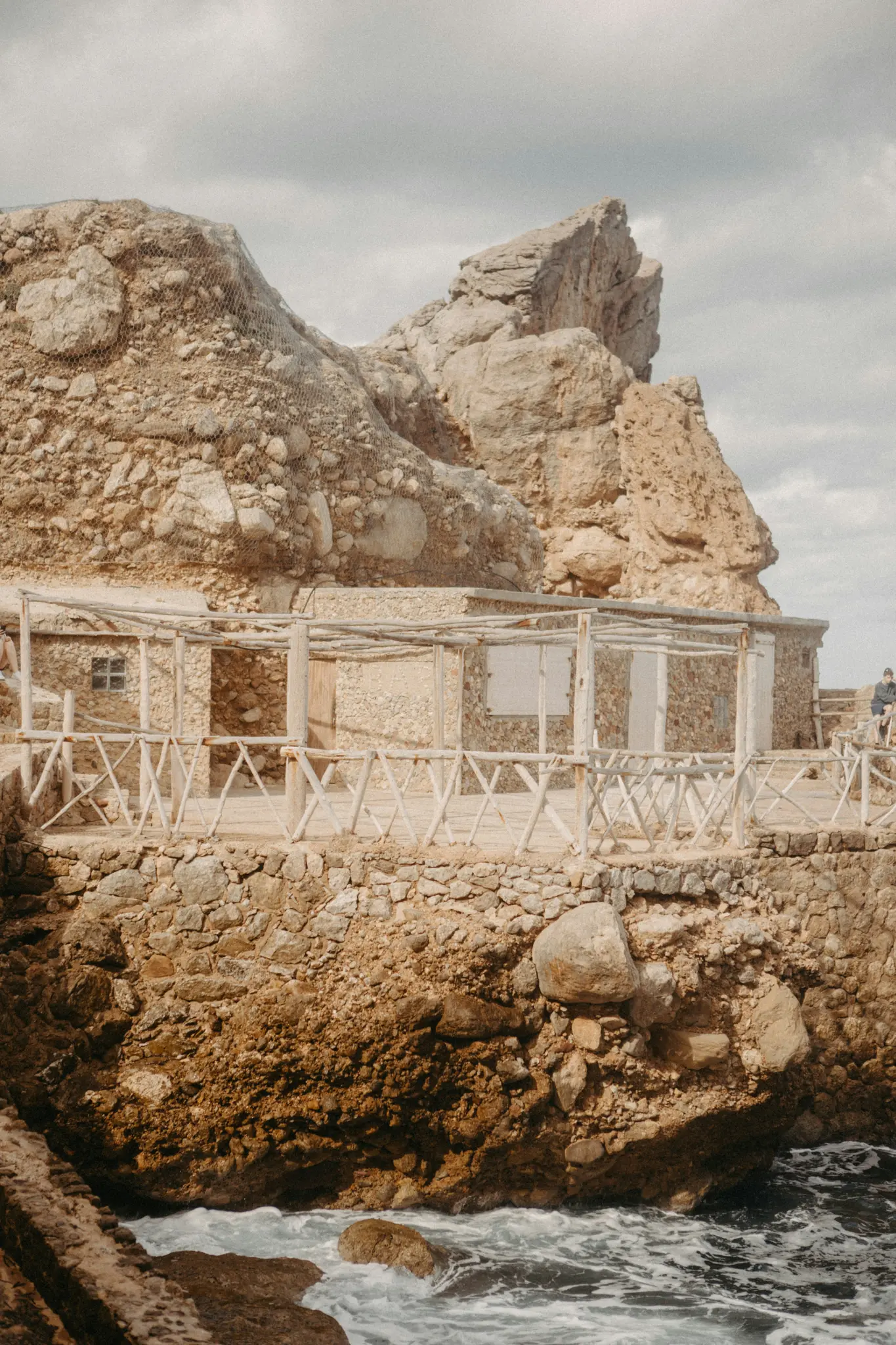 A rustic stone building with a wooden railing built into a rugged, rocky cliffside, with ocean waves crashing below.