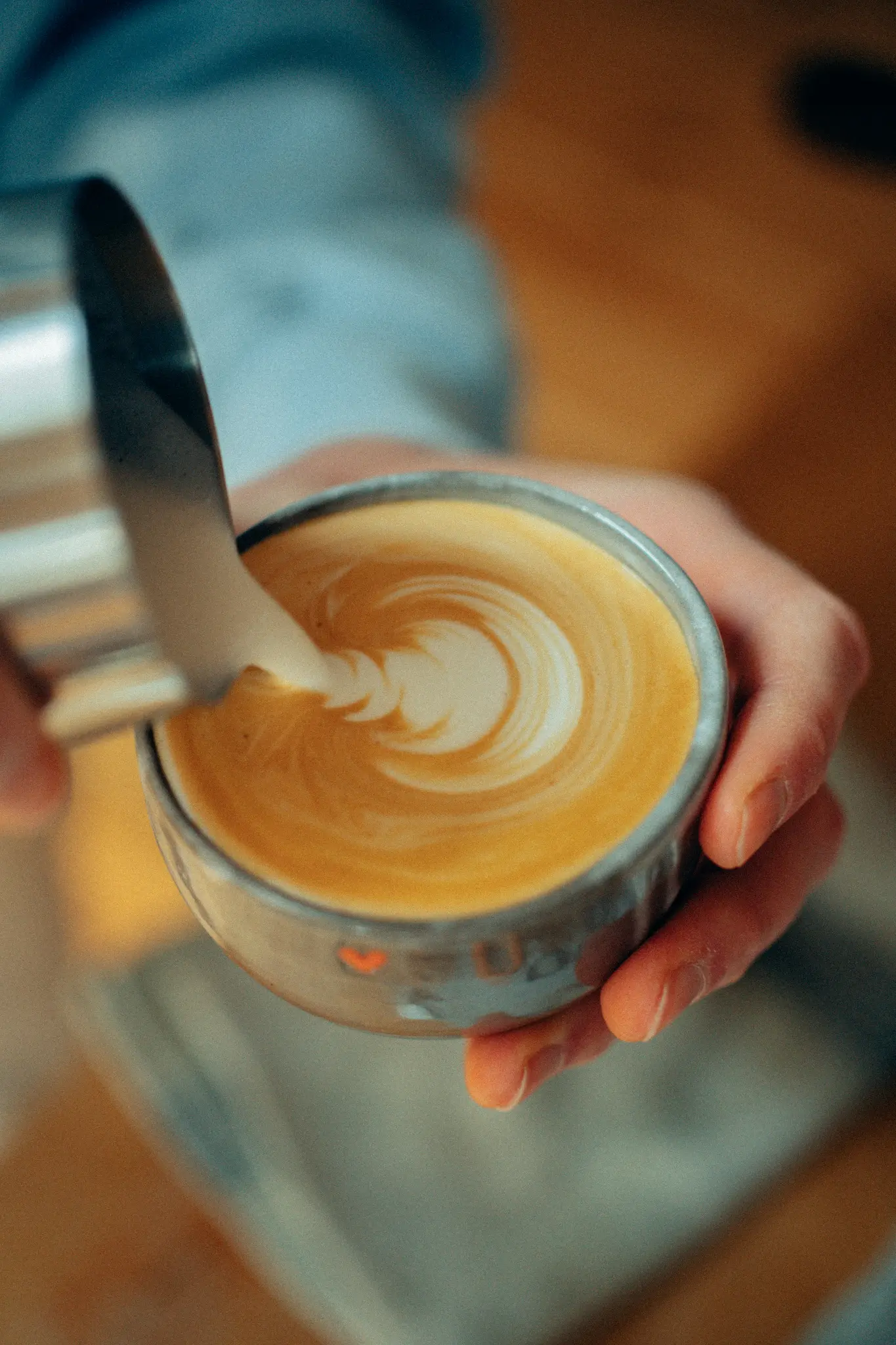 Close-up van handen die opgeschuimde melk uit een metalen schenkkan in een koffiekop gieten, waarbij een werveling van latte