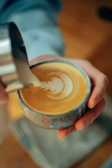 Close-up of hands pouring frothed milk from a metal pitcher into a cup of coffee, forming a swirl of latte art.