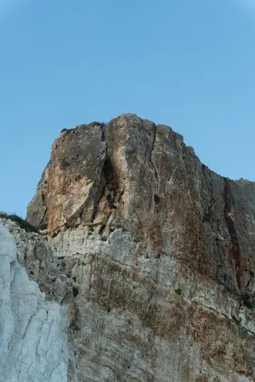 Obrovská, texturovaná skalní stěna se zvedá z levého dolního rohu do pravého horního a zobrazuje vrstvy hnědé, šedé a…
