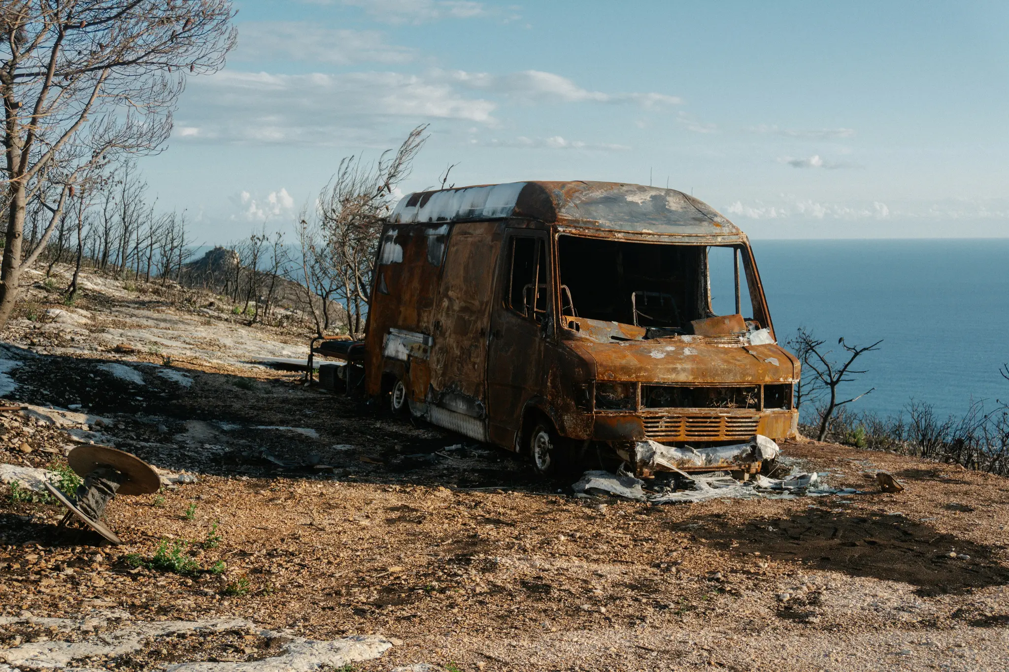 Ein stark verbrannter und verrosteter Campervan steht auf einem kargen, felsigen Hang. Das Fahrzeug ist schwarz und