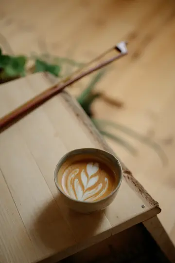 Keramický hrnek latte s detailním bílým designem latte art sedí na světlém dřevěném povrchu. Pozadí je…