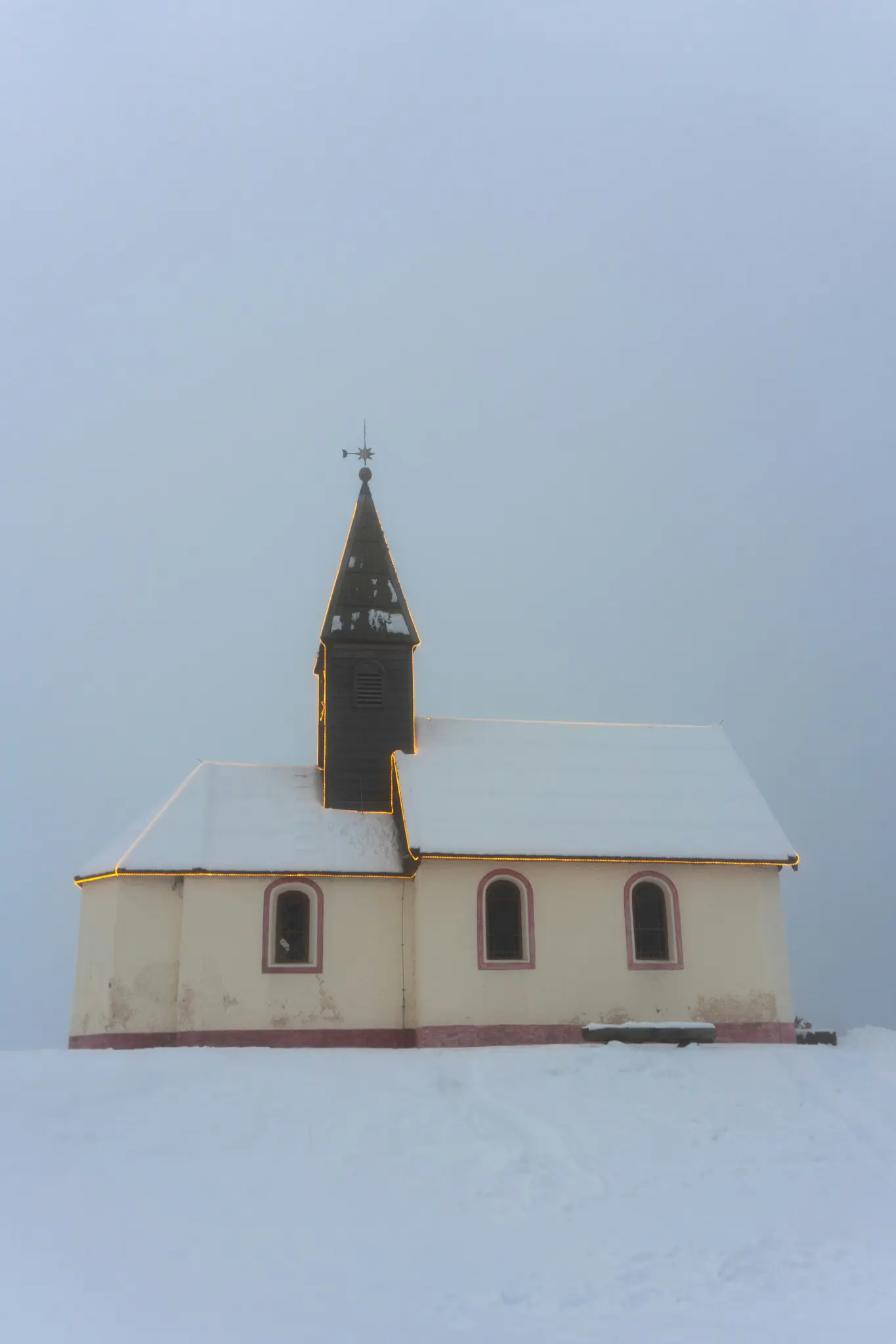 Een kleine, crèmekleurige kerk met een donkerbruine kerktoren staat in een veld met witte sneeuw onder een bewolkte hemel.
