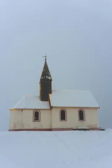 A small, cream-colored church with a dark brown steeple stands in a field of white snow under an overcast sky. Golden…