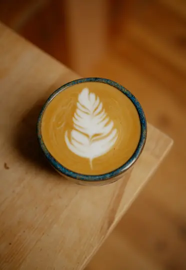 Detailní záběr shora na latte v keramickém šálku s modrým okrajem, s jemným bílým latte art ve tvaru listu na světlém pozadí