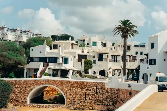 A picturesque village scene featuring numerous white buildings with green shutters and balconies, a stone arch bridge, and…