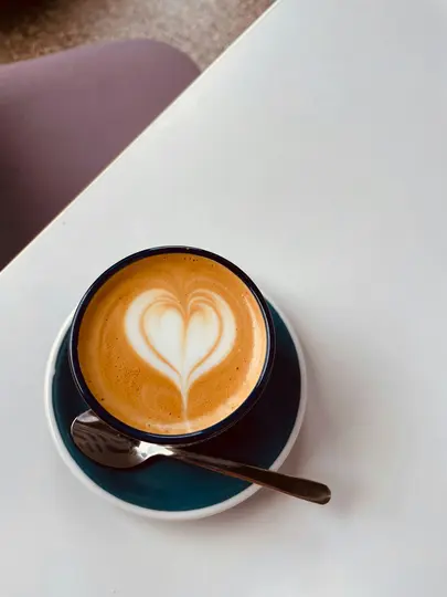 A top-down view of a latte with a heart design in the foam, served in a dark blue cup on a teal saucer with a spoon, all…