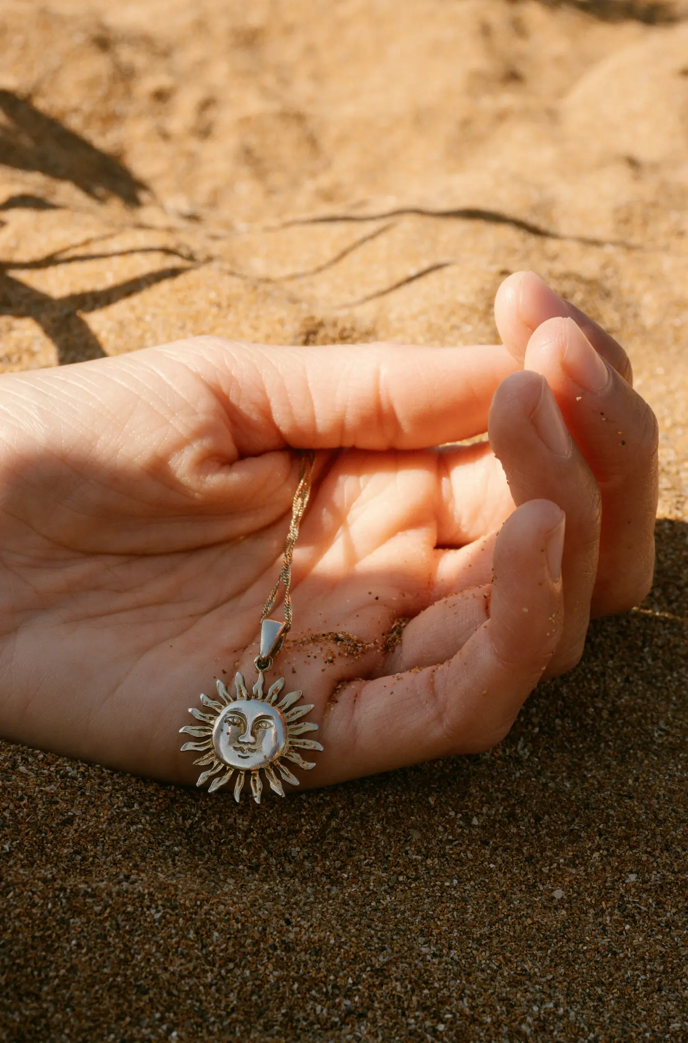 Eine Nahaufnahme einer Hand, die auf Sand ruht und einen silbernen Sonnenanhänger mit Gesicht hält, der an einer Goldkette