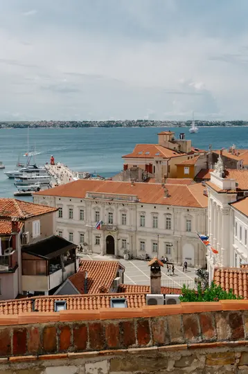 An elevated view of a coastal town featuring numerous buildings with orange-red terracotta roofs, a large white building…