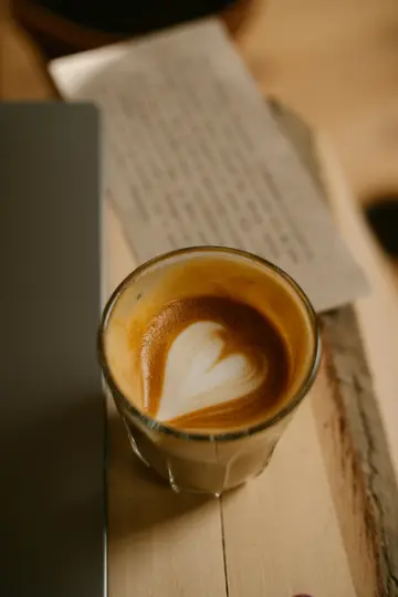 Un primo piano dall'alto di un bicchiere di latte con un disegno a cuore bianco nella schiuma, accanto a un laptop e a un
