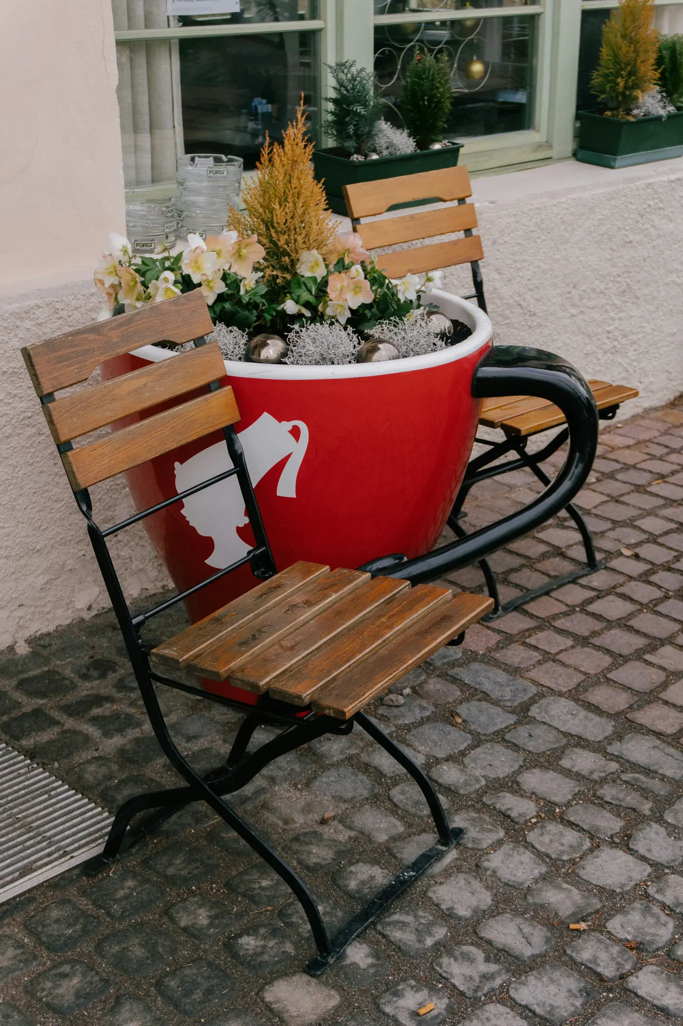 Een grote rode koffiekop plantenbak met een witte silhouet van een vrouwenhoofd en een kan, gevuld met planten en bloemen.