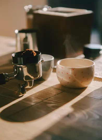 Un portafiltro con caffè macinato e una tazza bianca fumante con cuori rossi sono su un tavolo di legno, illuminati da una