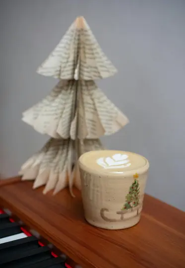 A ceramic cup of latte with a heart-shaped latte art sits on a wooden surface next to a Christmas tree made from folded…