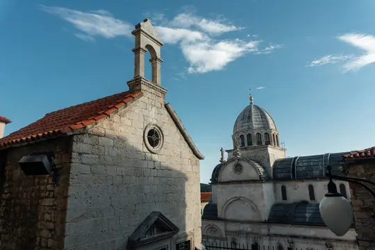 Kamena građevina sa zvonikom i krovom od crvenih pločica u prvom planu, djelomično u sjeni. Iza nje, velika kupola…