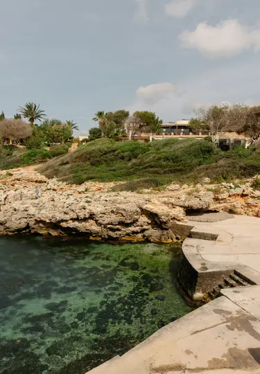 Μια γραφική θέα μιας βραχώδους όρμου με διαφανές πράσινο νερό, μια τσιμεντένια πλατφόρμα και μια πλαγιά καλυμμένη με πράσινη