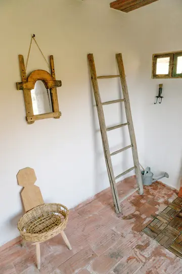 Rustikální místnost s bílými stěnami a terakotovou podlahou. Vysoký dřevěný žebřík se opírá o zeď vedle malého okna.
