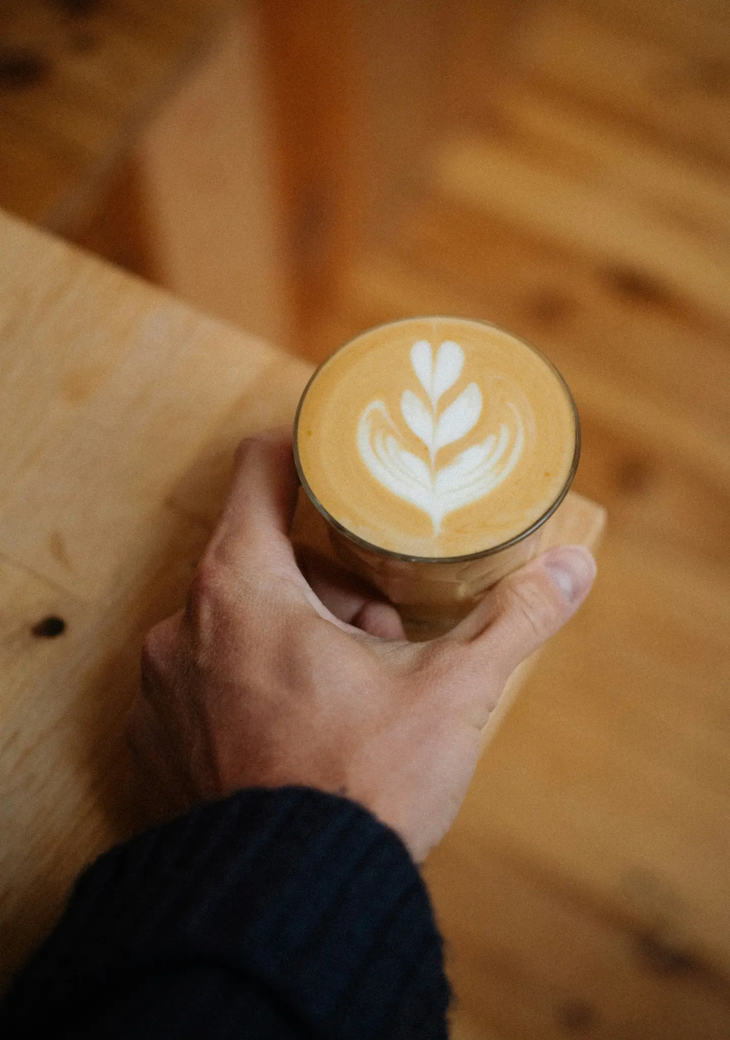 Ruka v tmavomodrom rukáve drží pohár latte so srdiečkom a listovým dizajnom.