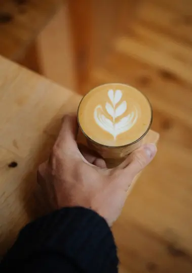 Una mano con una manica blu scuro tiene un bicchiere trasparente di latte con un disegno a forma di cuore e foglia.