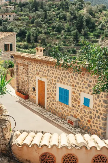 A stone house with a wooden door and two blue-shuttered windows stands on a paved street. Terraced hillsides with olive…