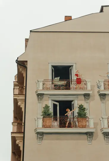 En bygningsfacade med malede balkoner og figurer. En kvinde i rødt holder en baby, et barn sigter med en bue, og en mand